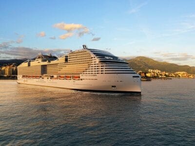 Una grande nave da crociera bianca MSC naviga vicino a una città costiera con maestose montagne sullo sfondo e il cielo azzurro del Mediterraneo.