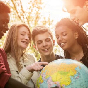 Cinque adolescenti sorridenti si trovano all'aperto alla luce del sole, riuniti intorno a un mappamondo. Una ragazza indica un luogo sul globo mentre le altre guardano con interesse. Sullo sfondo sono visibili alberi e luce del sole.