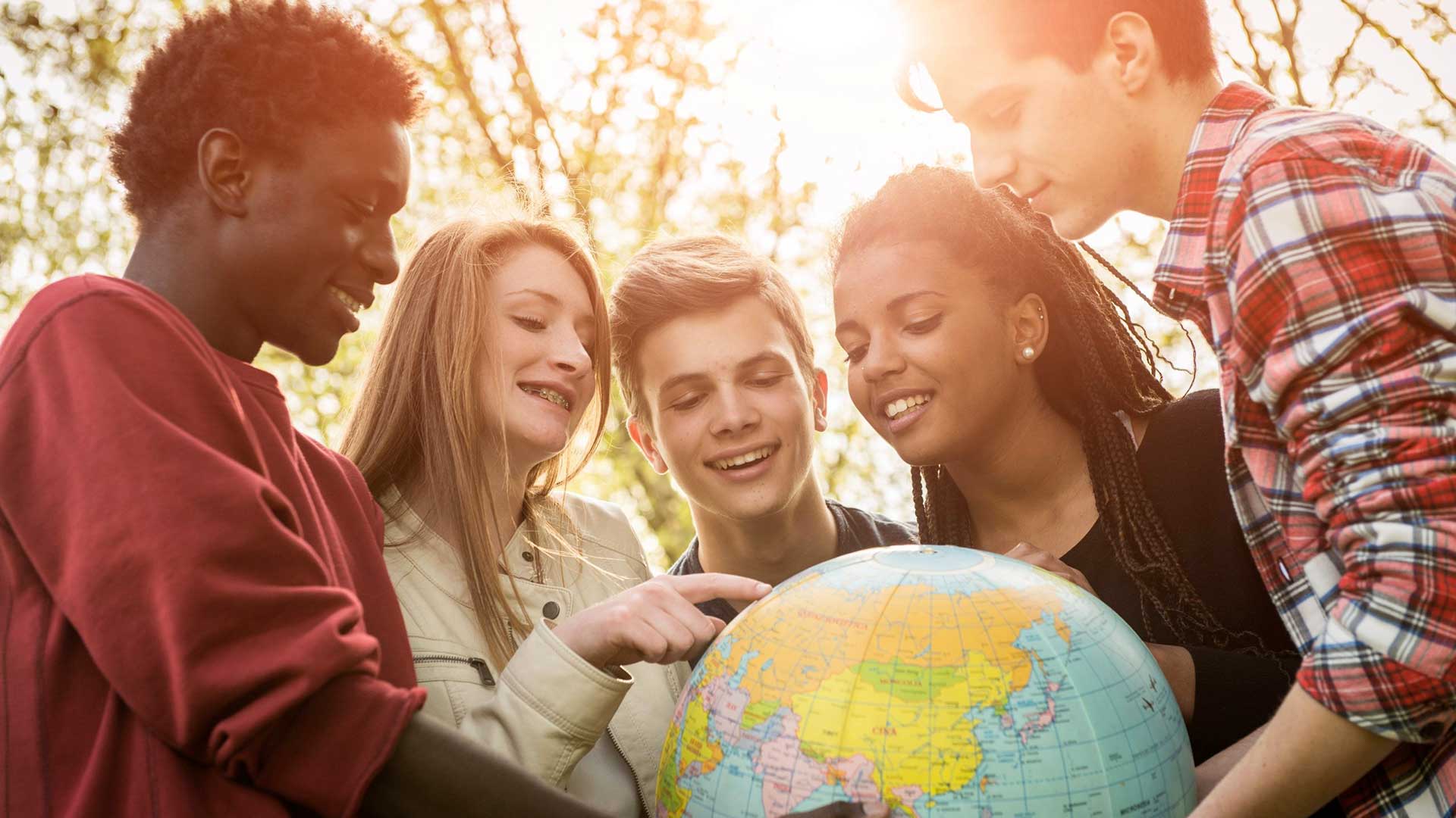 Cinque adolescenti sorridenti si trovano all'aperto alla luce del sole, riuniti intorno a un mappamondo. Una ragazza indica un luogo sul globo mentre le altre guardano con interesse. Sullo sfondo sono visibili alberi e luce del sole.