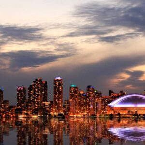 Lo skyline di Toronto al tramonto con la CN Tower e il Rogers Centre illuminati, gli edifici della città che si riflettono nell'acqua sotto un cielo parzialmente nuvoloso.