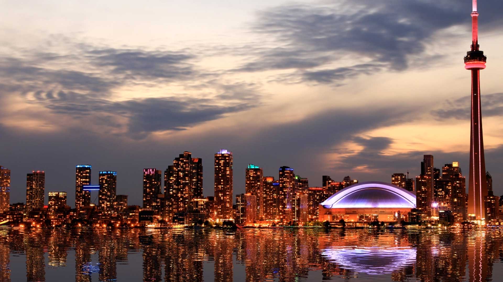 Lo skyline di Toronto al tramonto con la CN Tower e il Rogers Centre illuminati, gli edifici della città che si riflettono nell'acqua sotto un cielo parzialmente nuvoloso.