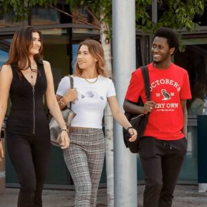 Quattro giovani adulti camminano e parlano fuori da un edificio con la scritta "Woodla... Boarding Ho...". Tre sono in abiti casual e sorridono, mentre uno sullo sfondo cammina da solo. Alle loro spalle sono visibili alberi e grandi finestre.