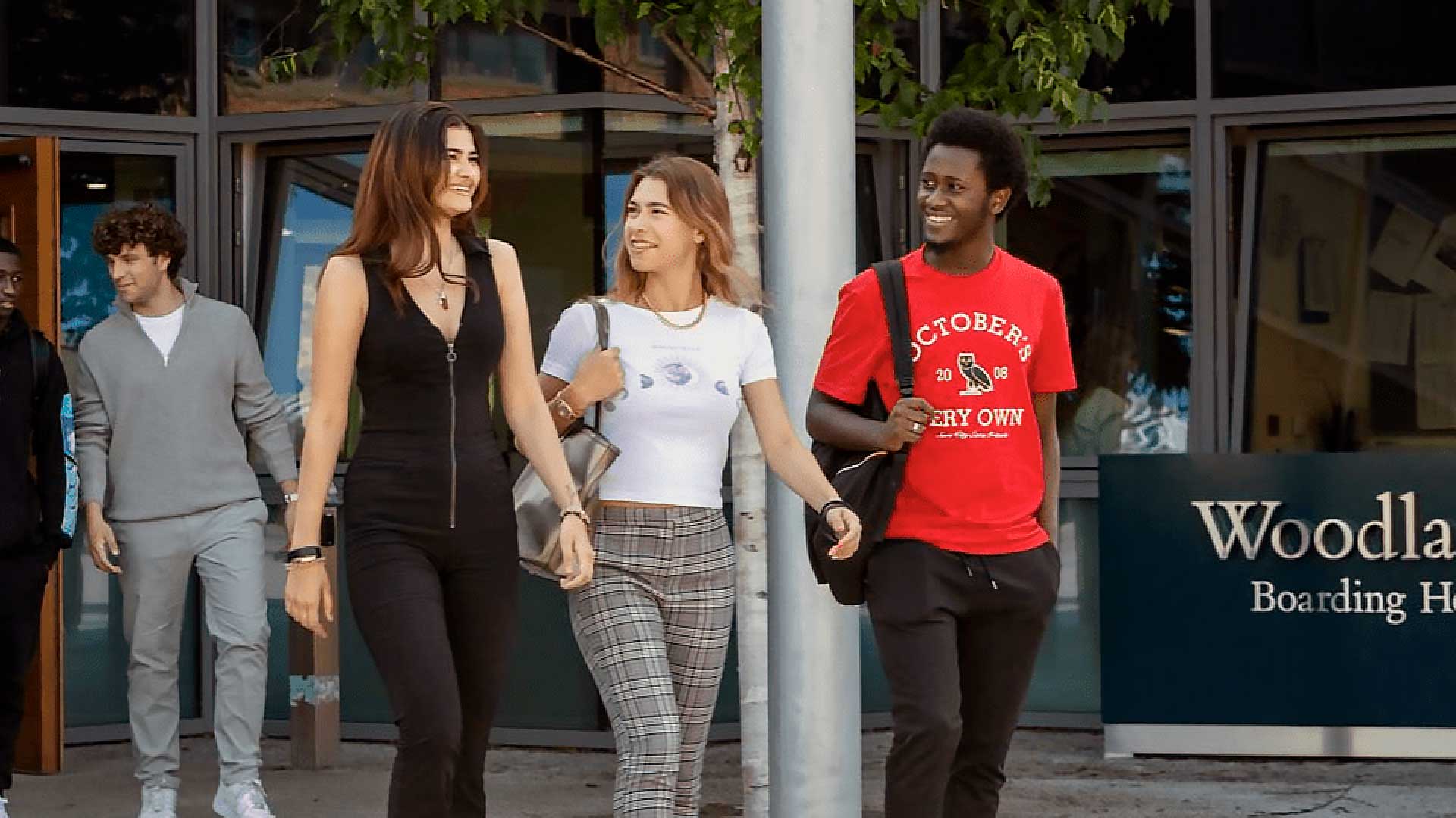 Quattro giovani adulti camminano e parlano fuori da un edificio con la scritta "Woodla... Boarding Ho...". Tre sono in abiti casual e sorridono, mentre uno sullo sfondo cammina da solo. Alle loro spalle sono visibili alberi e grandi finestre.