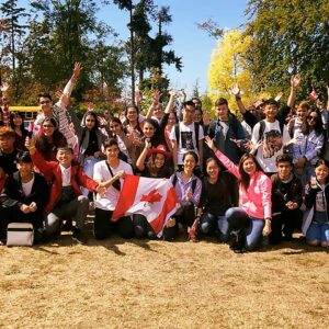 Un folto gruppo di giovani posa all'aperto in una giornata di sole, alcuni dei quali tengono o indossano bandiere canadesi. Sorridono, fanno il tifo e sono vestiti in modo casual, con alberi e scuolabus sullo sfondo.
