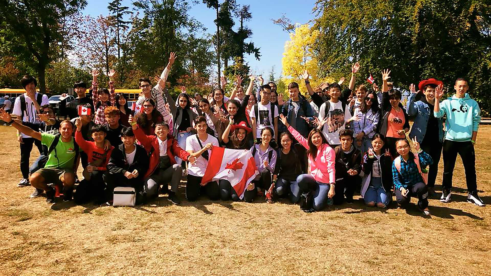 Un folto gruppo di giovani posa all'aperto in una giornata di sole, alcuni dei quali tengono o indossano bandiere canadesi. Sorridono, fanno il tifo e sono vestiti in modo casual, con alberi e scuolabus sullo sfondo.