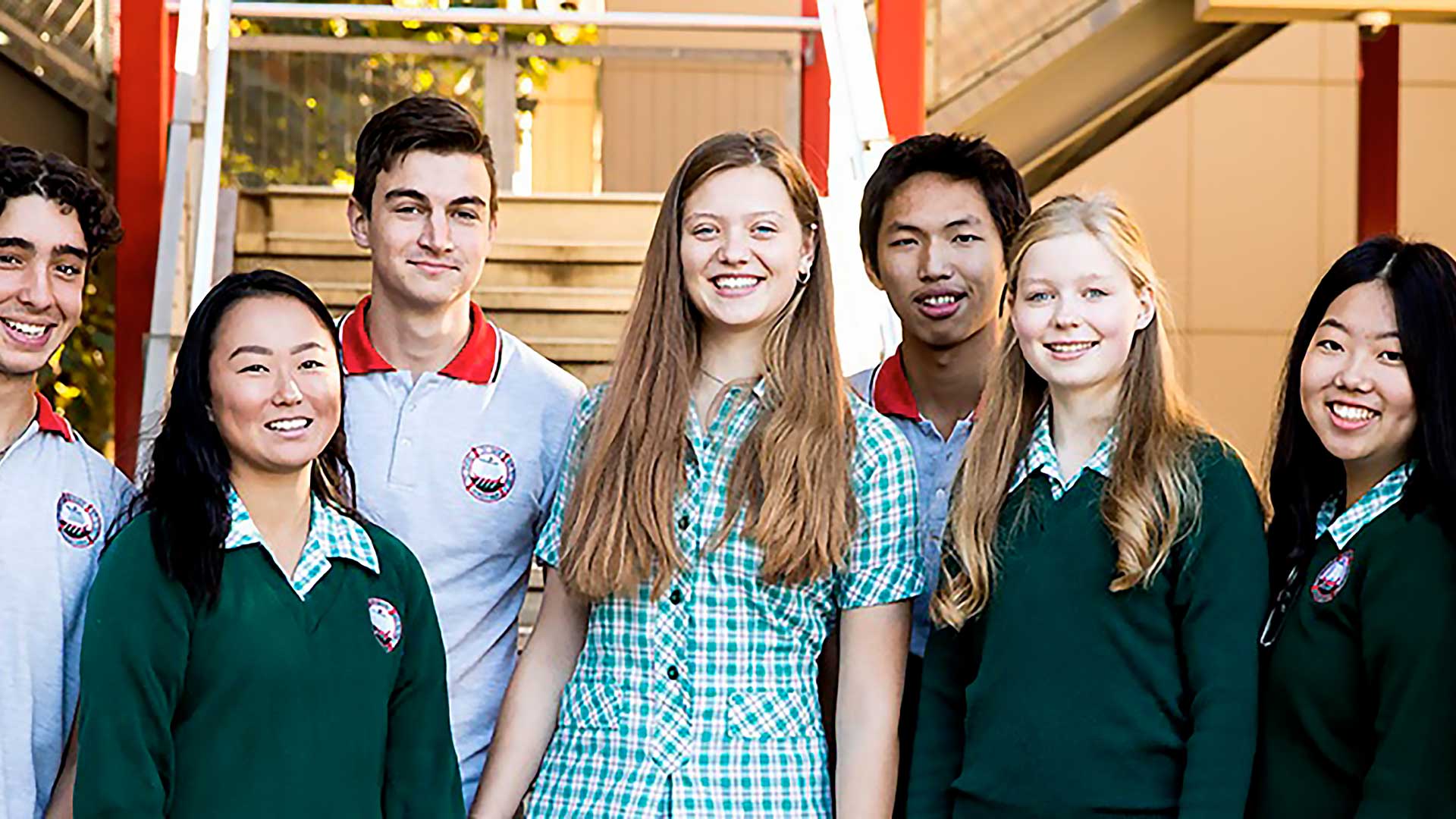 Un gruppo di sette studenti liceali sorridenti, alcuni con maglioni verdi e altri con polo grigie, in piedi all'aperto vicino alle scale.