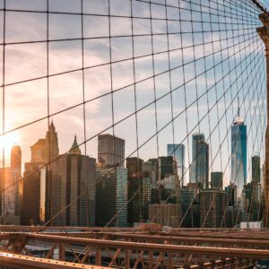 Veduta dello skyline di New York al tramonto, visto attraverso i cavi del ponte di Brooklyn, con la luce del sole che illumina gli edifici e la struttura del ponte.