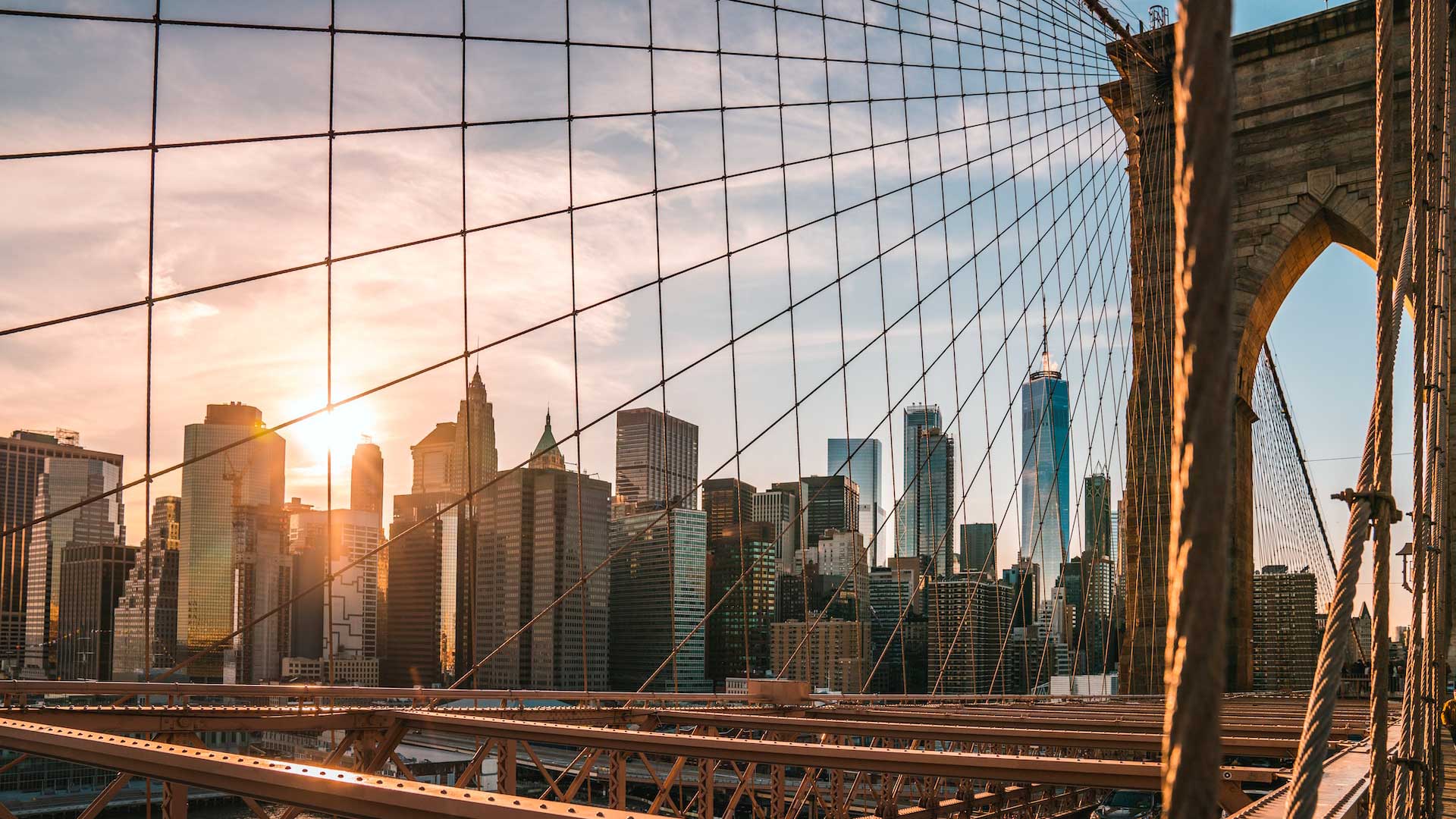 Veduta dello skyline di New York al tramonto, visto attraverso i cavi del ponte di Brooklyn, con la luce del sole che illumina gli edifici e la struttura del ponte.