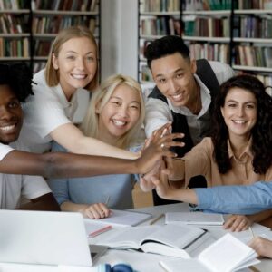 Un gruppo eterogeneo di sei giovani adulti seduti a un tavolo in una biblioteca, che sorridono e uniscono le mani al centro su libri aperti e un computer portatile, con gli scaffali sullo sfondo.
