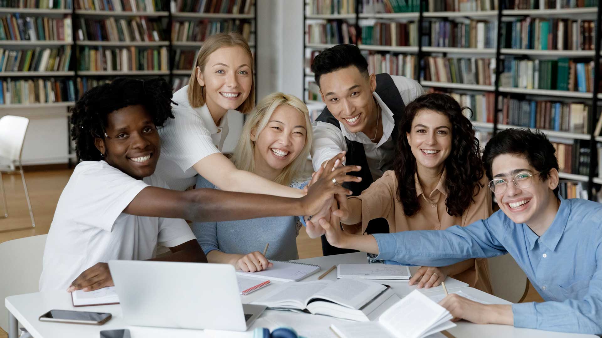 Un gruppo eterogeneo di sei giovani adulti seduti a un tavolo in una biblioteca, che sorridono e uniscono le mani al centro su libri aperti e un computer portatile, con gli scaffali sullo sfondo.