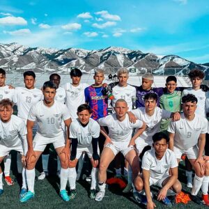 Una squadra di calcio di giovani uomini in uniforme bianca posa insieme su un campo con montagne innevate e un cielo azzurro sullo sfondo. Alcuni giocatori sono in piedi, altri in ginocchio e tutti guardano verso la telecamera.