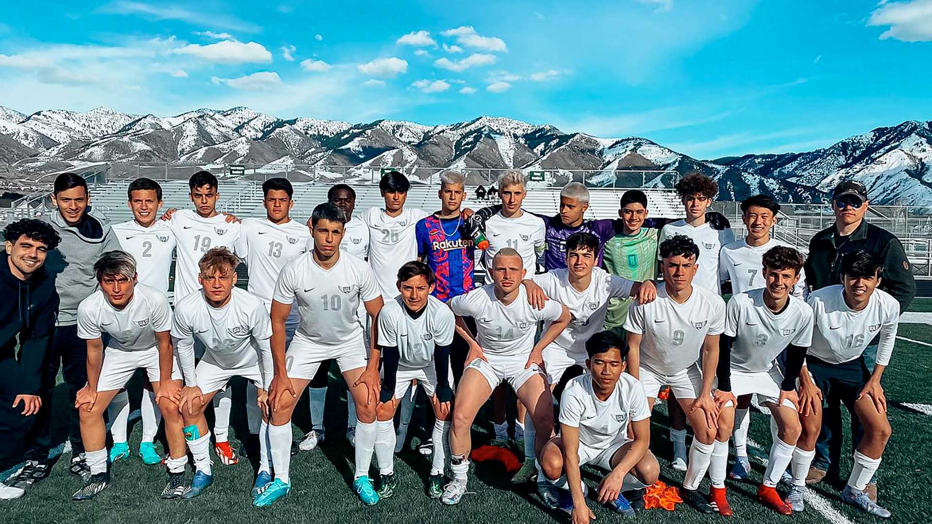 Una squadra di calcio di giovani uomini in uniforme bianca posa insieme su un campo con montagne innevate e un cielo azzurro sullo sfondo. Alcuni giocatori sono in piedi, altri in ginocchio e tutti guardano verso la telecamera.