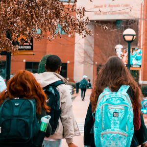Cinque studenti con lo zaino in spalla camminano insieme in un campus universitario verso un edificio di mattoni, circondato da alberi spogli e dalla segnaletica del campus, in una giornata nuvolosa.