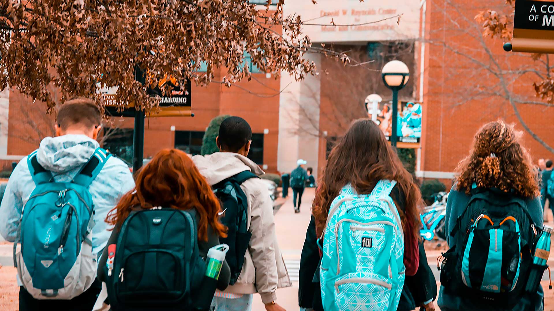 Cinque studenti con lo zaino in spalla camminano insieme in un campus universitario verso un edificio di mattoni, circondato da alberi spogli e dalla segnaletica del campus, in una giornata nuvolosa.