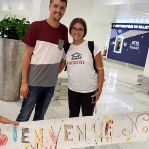 Una famiglia sorridente di quattro persone si trova in un aeroporto con in mano un cartello colorato fatto a mano con la scritta "Bienvenue Noel". Due bambini, un maschio e una femmina, tengono il cartello, mentre i genitori stanno dietro di loro.