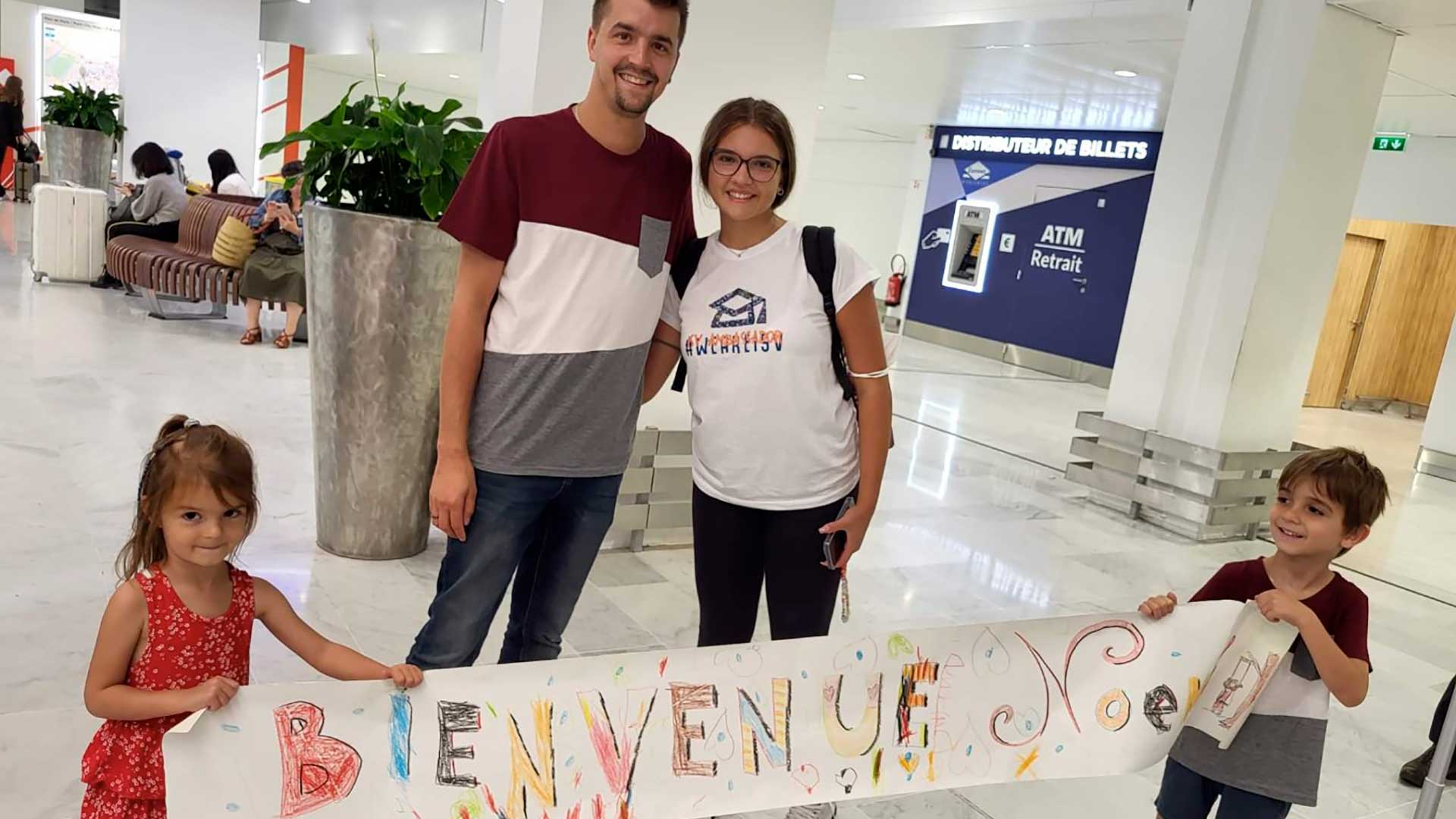 Una famiglia sorridente di quattro persone si trova in un aeroporto con in mano un cartello colorato fatto a mano con la scritta "Bienvenue Noel". Due bambini, un maschio e una femmina, tengono il cartello, mentre i genitori stanno dietro di loro.