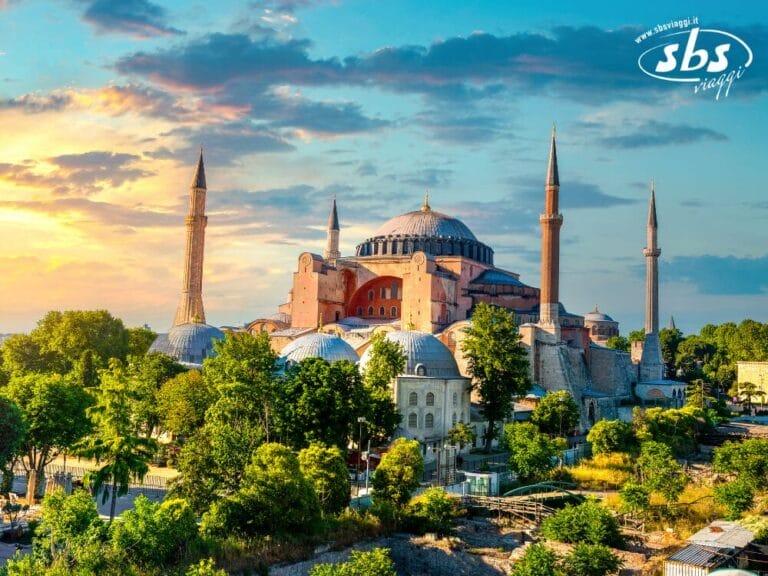 L'immagine mostra Hagia Sophia, un sito architettonico storico a Istanbul, Turchia, circondato dal verde con un cielo parzialmente nuvoloso al tramonto. Considera di aggiungere questa vista mozzafiato al tuo tour del 2025 in Turchia per un'esperienza indimenticabile.