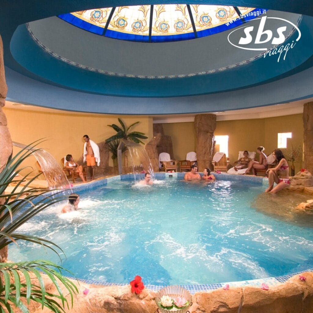 Persone che si rilassano e si divertono in una piscina coperta del Caribbean World Resort, Soma Bay. La scena comprende formazioni rocciose decorative, un soffitto a cupola in vetro colorato, giochi d'acqua e piante tropicali.