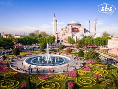 Vista aerea di Hagia Sophia con i suoi minareti, circondata da un giardino con una fontana centrale, aiuole colorate e sentieri pieni di gente. Vivi questa vista mozzafiato nel Tour Eleganza Della Turchia 2025.