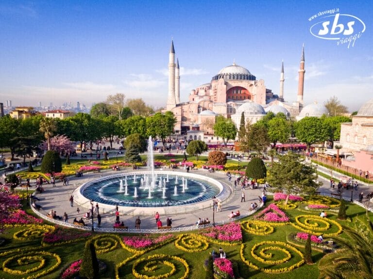 Vista aerea di Hagia Sophia con i suoi minareti, circondata da un giardino con una fontana centrale, aiuole colorate e sentieri pieni di gente. Vivi questa vista mozzafiato nel Tour Eleganza Della Turchia 2025.