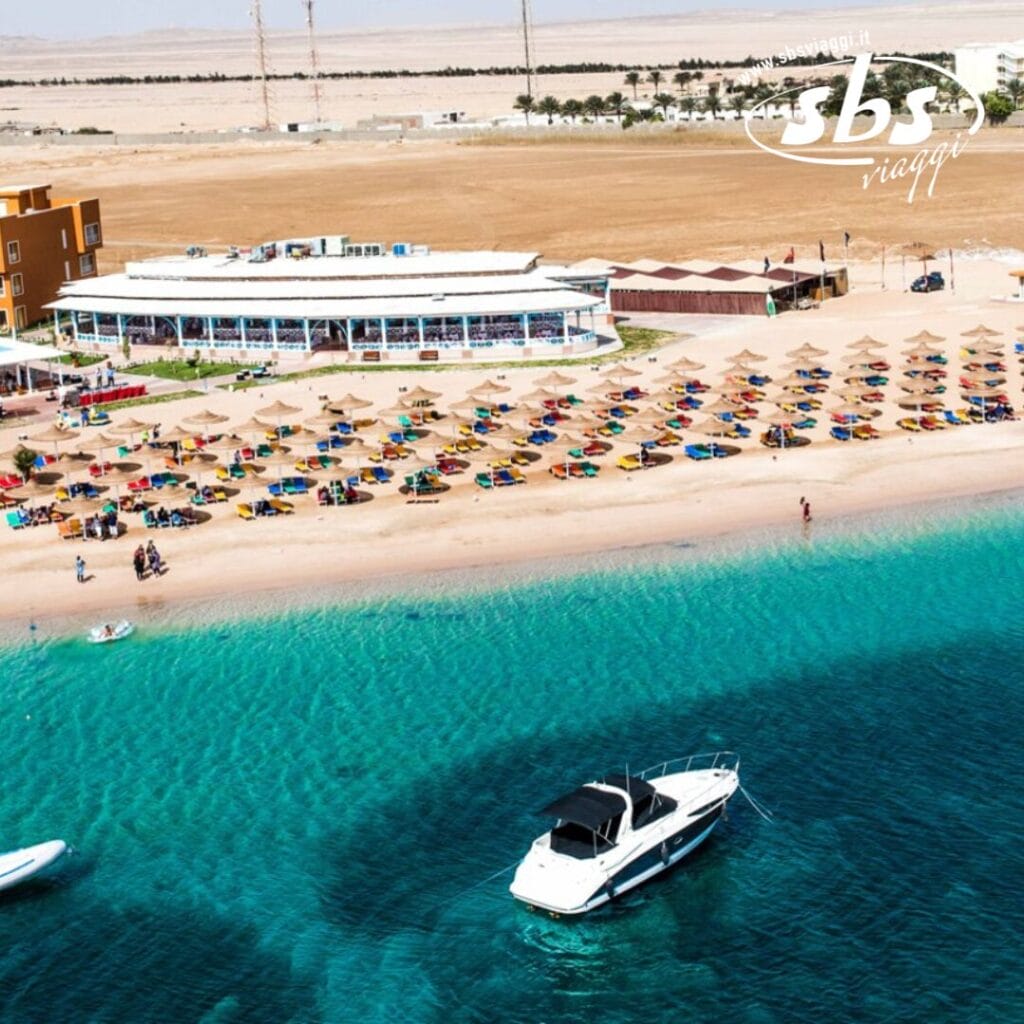 Vista aerea di Soma Bay con un resort sulla spiaggia con file di ombrelloni colorati e lettini, un edificio bianco e una barca ancorata in acque azzurre e limpide. Sullo sfondo sono visibili terreni marroni e strutture secondarie.