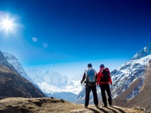 Due escursionisti con zaini e bastoncini da trekking sono in piedi su un terreno roccioso, con vista sulle montagne innevate sotto un cielo azzurro e terso con il sole che splende luminoso. La scena cattura un momento di Bozza automatica che sembra quasi surreale nella sua bellezza naturale.