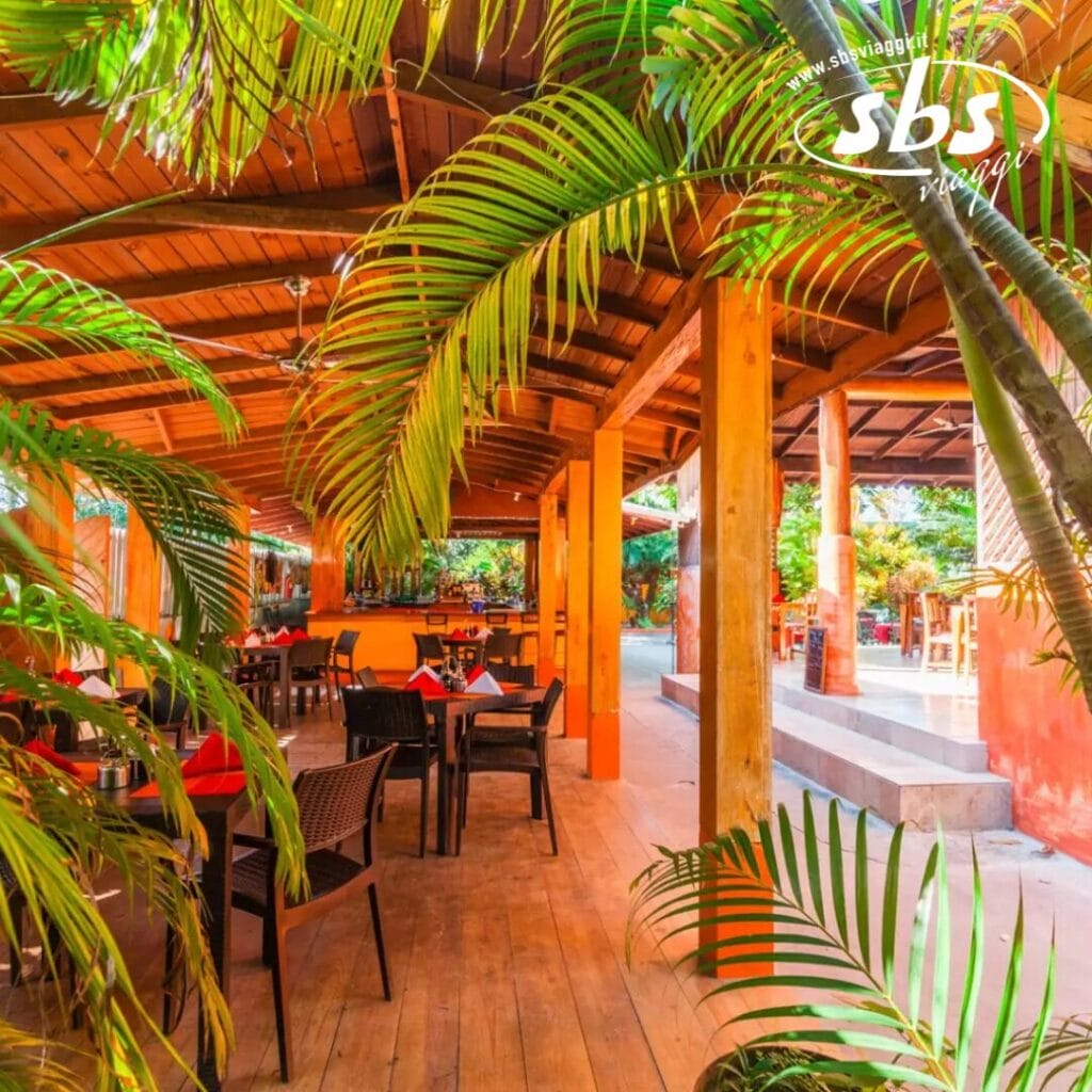 L'area salotto all'aperto del Sunrise Club è dotata di tavoli e sedie in legno, circondati da una vegetazione lussureggiante sotto un affascinante tetto in legno. La luce naturale inonda lo spazio, creando un'atmosfera invitante, perfetta per le riunioni mattutine o per i pomeriggi di relax.