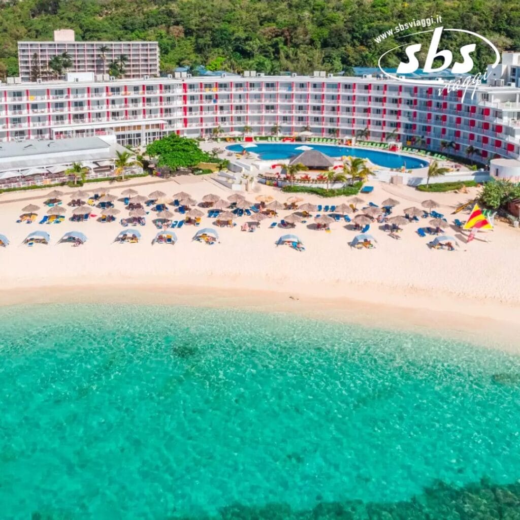 Vista aerea di un resort sulla spiaggia, caratterizzato da un hotel in stile bozza automatica, una piscina scintillante e file di ombrelloni e sedie a sdraio sulla spiaggia sabbiosa, il tutto sullo sfondo di un'acqua turchese e limpida.