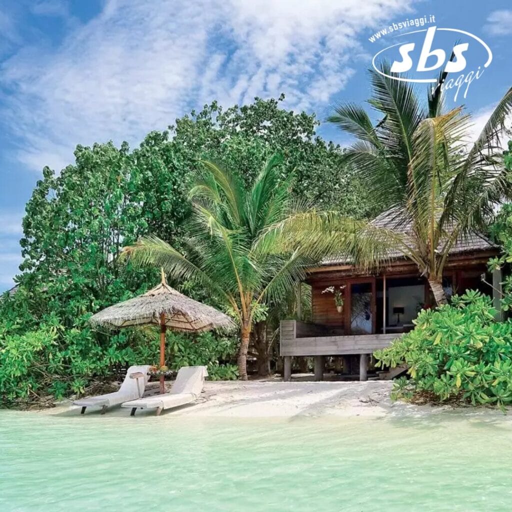 Scena di spiaggia tropicale con due sedie a sdraio sotto un ombrellone di paglia, vicino a una capanna di legno circondata da una vegetazione lussureggiante e palme, che ricorda l'atmosfera serena del Gangehi Island Resort.