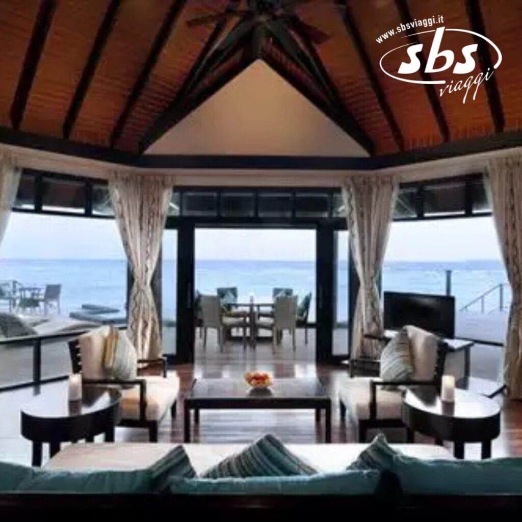Un lussuoso soggiorno all'Iru Fushi con ampie finestre si affaccia sull'oceano. La stanza, parte del resort Sun Siyam alle Maldive, è dotata di mobili eleganti e di una zona pranzo, che fonde perfettamente comfort e stile.