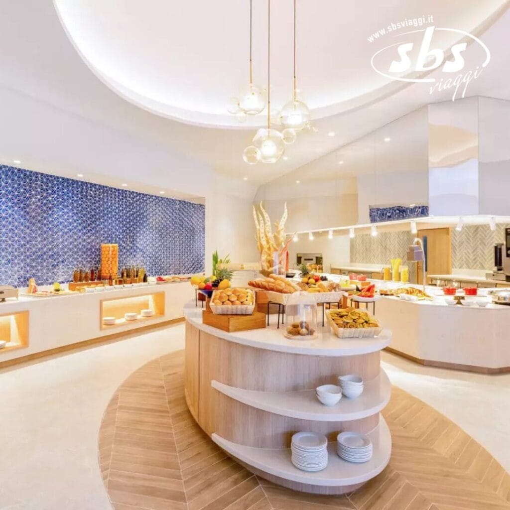 Un'area buffet ben illuminata con un'isola centrale che espone una varietà di cibi nello stile di Nova Maldives. Lo sfondo presenta una parete con motivi blu tipici dello straordinario design di Vakarufalhi Island e ulteriori postazioni di ristorazione che ricordano un resort di lusso delle Maldive.