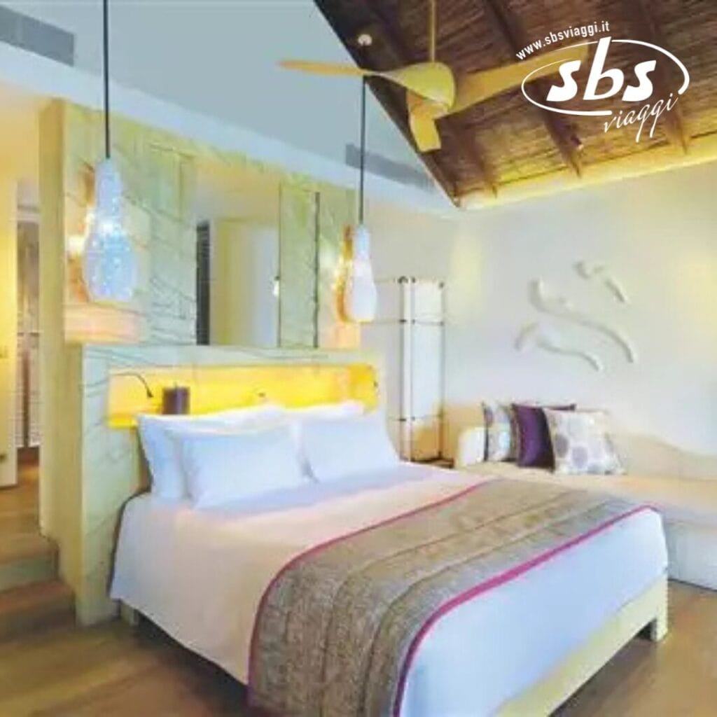 Una camera d'albergo accogliente al Constance Moofushi alle Maldive, con un letto grande, due lampade a sospensione e un ventilatore a soffitto in legno. Il design è moderno con una tavolozza di colori caldi e una discreta arte murale. Godetevi il lusso di un bagno parzialmente aperto in questa serena fuga sull'isola.