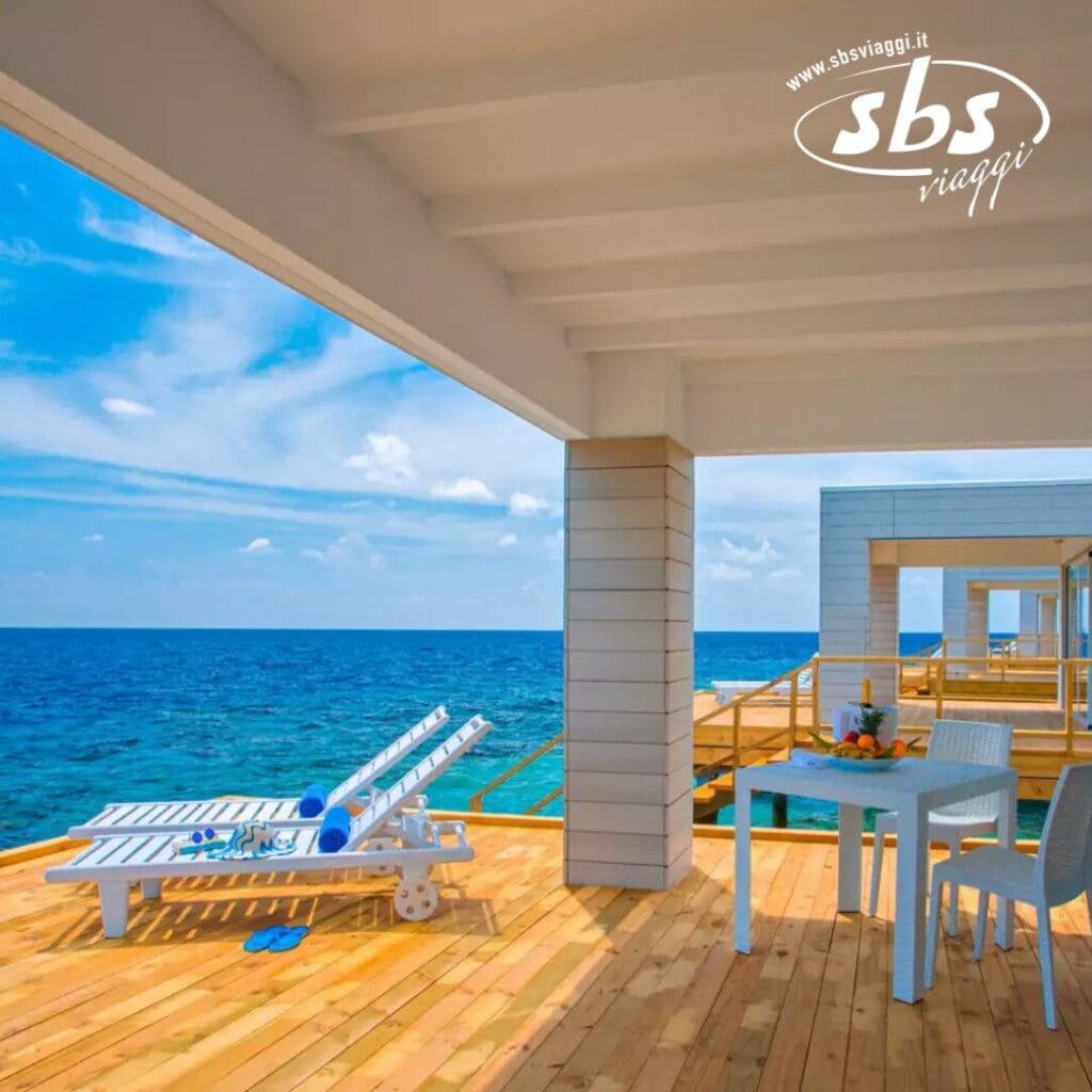 Deck in legno con sedie a sdraio e un tavolo apparecchiato, con vista sull'oceano blu brillante. Un cielo soleggiato con nuvole sparse esalta la scena tranquilla. Il logo Bozza automatica è elegantemente posizionato nell'angolo in alto a destra, aggiungendo un tocco di raffinatezza.