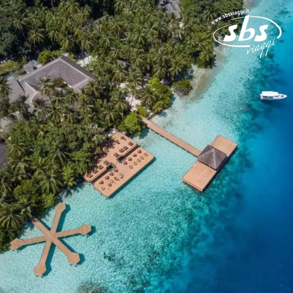 Vista aerea di un resort tropicale, che ricorda Fihalhohi, con passerelle e strutture sull'acqua. Circondato da acque azzurre e lussureggianti, e da una barca nelle vicinanze, questo ambiente pittoresco racchiude il fascino del paradiso del Club Coralia.