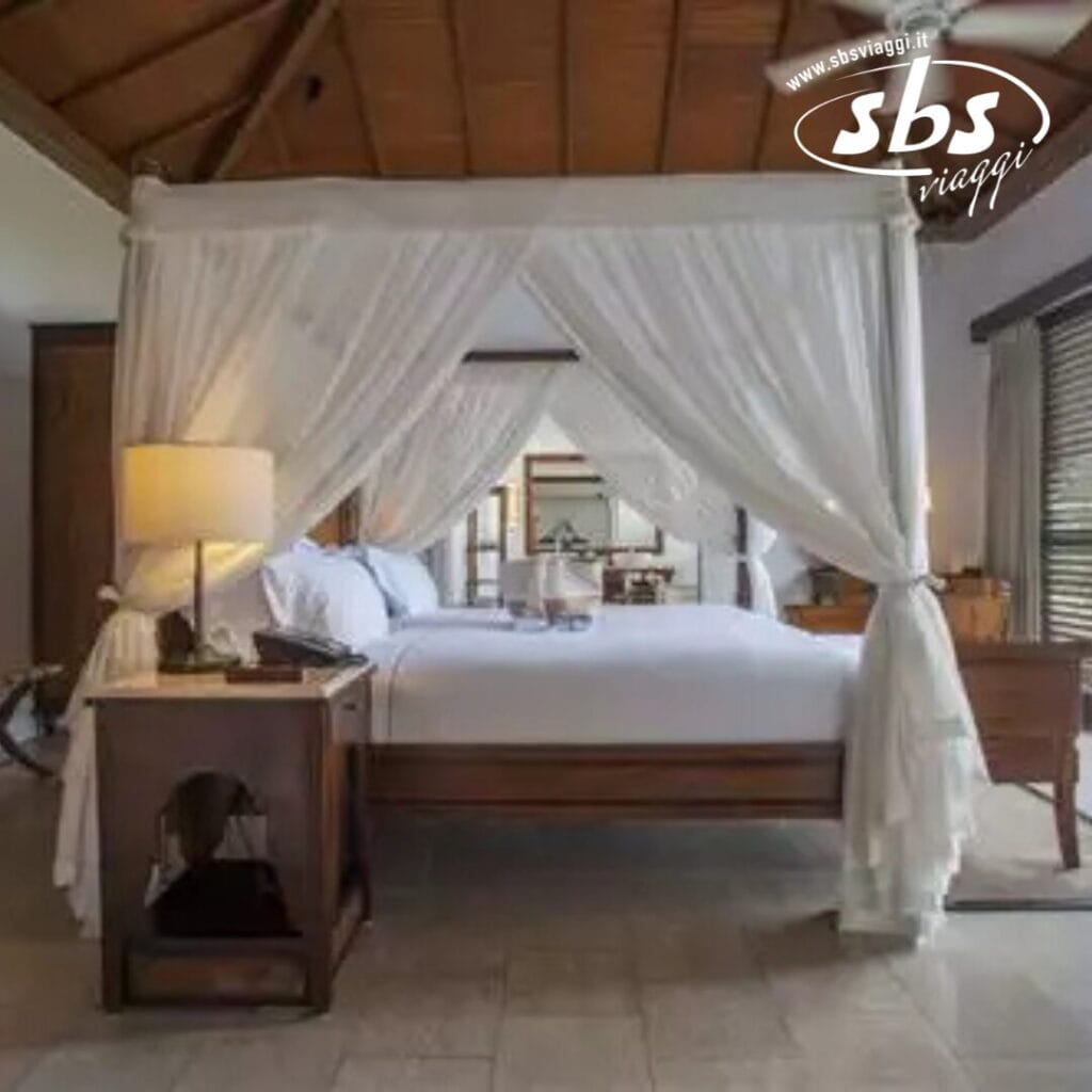 Una camera da letto in un resort tropicale con un letto a baldacchino drappeggiato in tende bianche, un comodino con una lampada e una grande finestra che mostra il logo SBS Viaggi. Perfetto per chiunque cerchi il lusso del Residence Zanzibar.