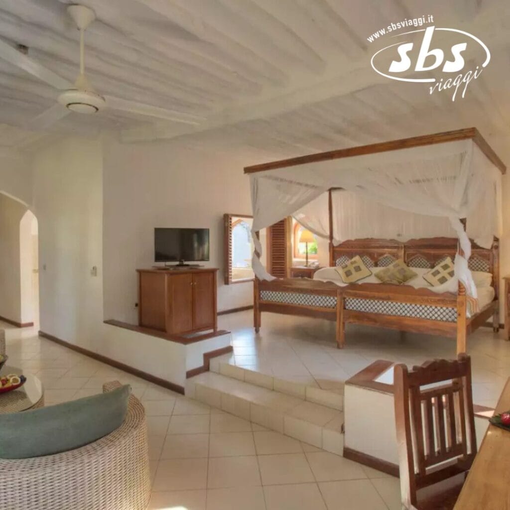 Una camera da letto accogliente al Bluebay Beach Resort dispone di un letto a baldacchino, una TV su un supporto di legno e una sedia di vimini. Con pareti chiare e pavimento piastrellato, la stanza trasuda tranquillità. Un arco aperto offre un accesso senza soluzione di continuità a un altro spazio, riecheggiando l'atmosfera rilassante della spa del resort.
