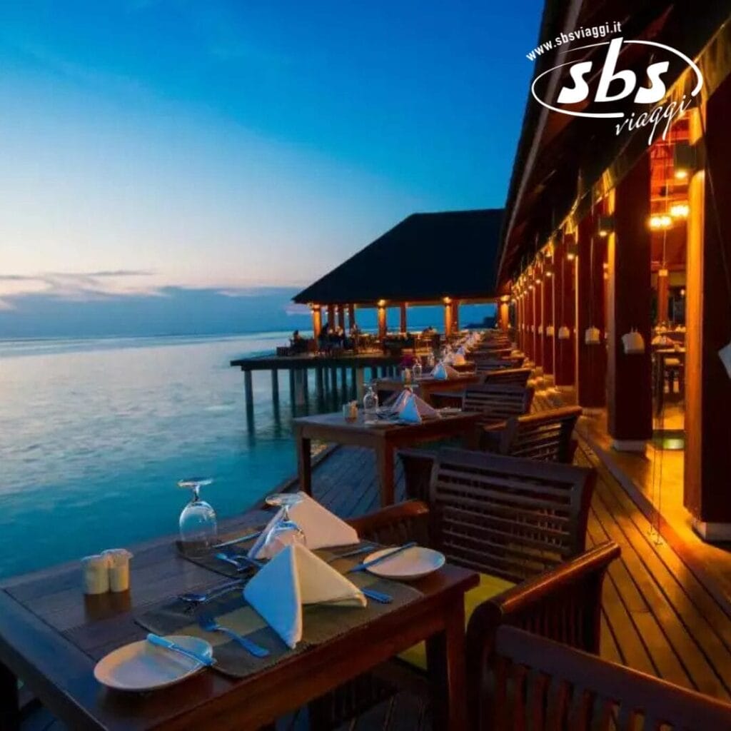 I tavoli da pranzo allestiti su una terrazza in legno con vista sull'oceano al tramonto evocano l'essenza di un'idilliaca serata estiva, con un affascinante edificio in legno che si estende sull'acqua, catturando perfettamente l'atmosfera rilassata dell'isola.