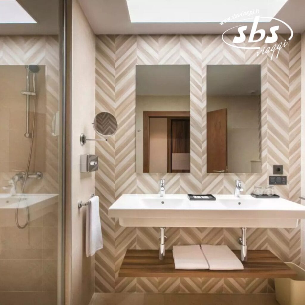 Questo moderno bagno del Riu Jambo Hotel è dotato di un elegante doppio lavabo, un grande specchio e pareti con motivi geometrici alla moda. Sulla sinistra, troverete una spaziosa cabina doccia che aggiunge fascino contemporaneo.