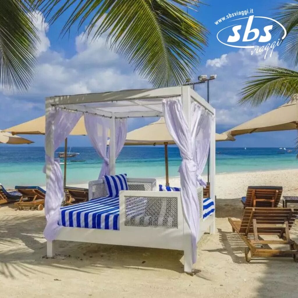 Scena di spiaggia in un resort sulla spiaggia di Zanzibar con un letto a baldacchino bianco drappeggiato in eleganti tende, cuscini a righe blu e circondato da palme, ombrelloni e sedie a sdraio. L'incantevole vista sull'oceano del Seaclub completa il cielo parzialmente nuvoloso sullo sfondo.