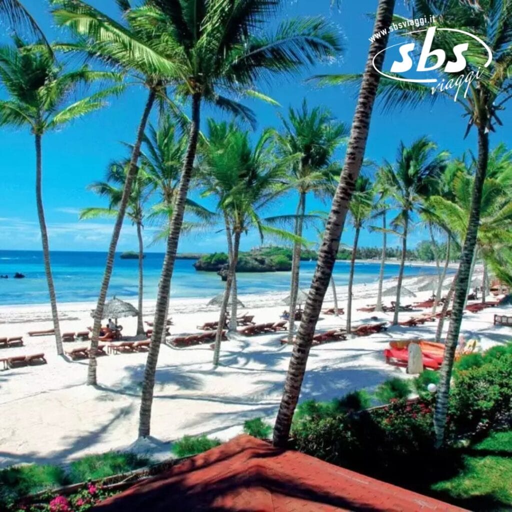 Scena di spiaggia con palme, sedie a sdraio e il mare azzurro e limpido di Crystal Bay sotto un cielo luminoso. Le strutture con i tetti rossi del resort Veraclub sono parzialmente visibili.