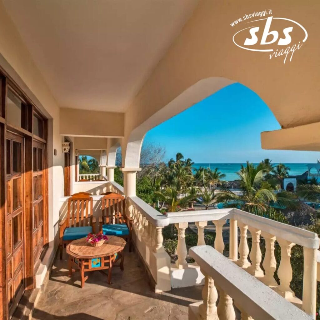 Un tranquillo balcone del Jumbo Resort, arredato con mobili in legno, offre una vista mozzafiato sul giardino tropicale e sull'oceano, sotto un limpido cielo azzurro.