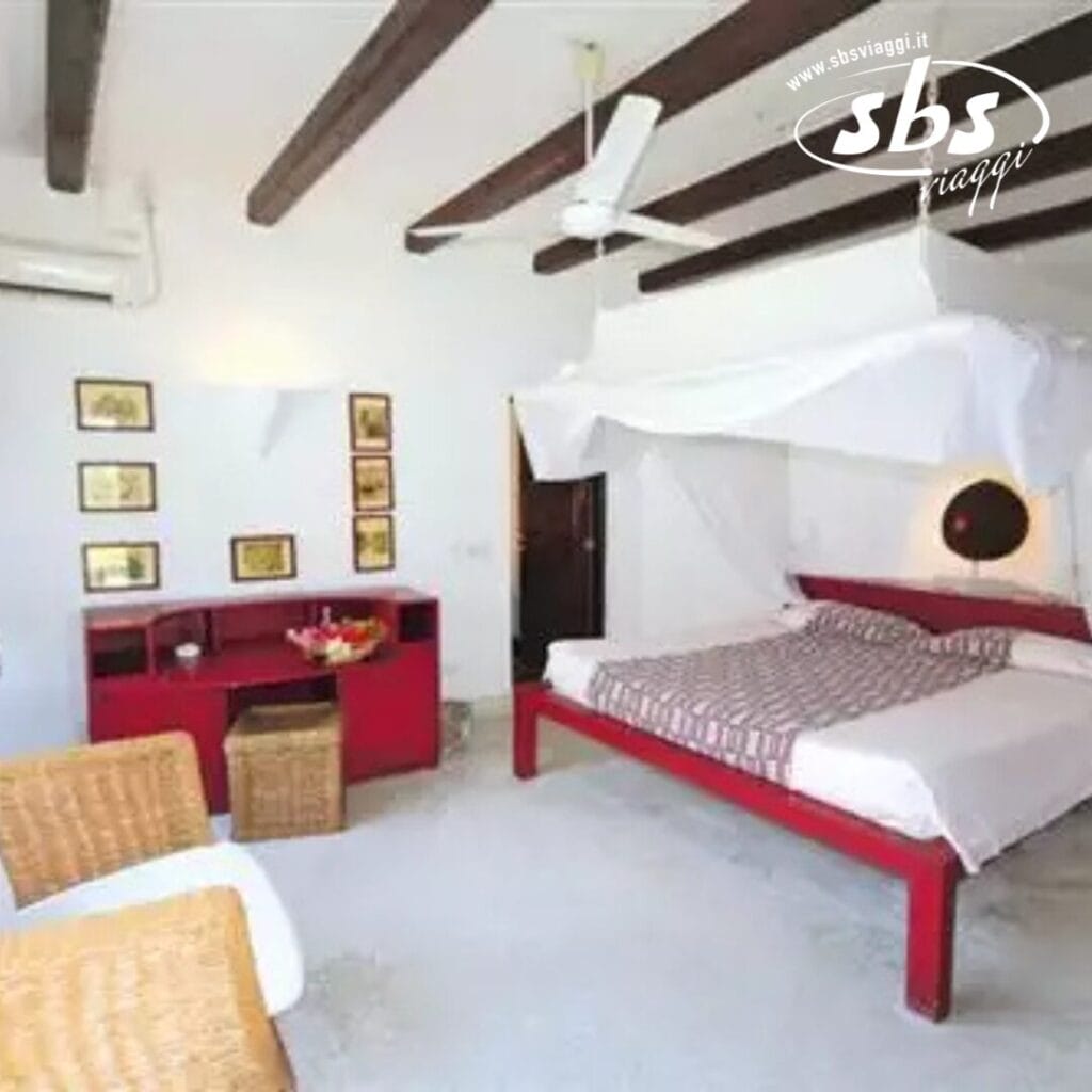 Una camera da letto con un letto rosso, un baldacchino bianco, una scaffalatura rossa, sedie in vimini e quadri incorniciati sul muro. Sono visibili il ventilatore a soffitto e l'unità di aria condizionata.