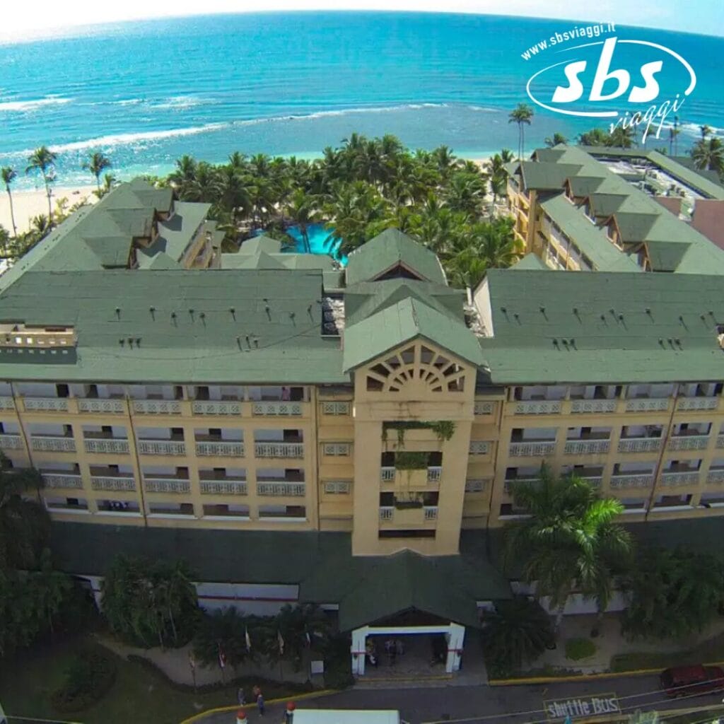 Una vista aerea del Coral Costa Caribe rivela un hotel sulla spiaggia con tetti verdi e lussureggianti, incastonato tra palme ondeggianti. Il vivace Beach Resort offre viste mozzafiato con l'oceano che si estende serenamente sullo sfondo.