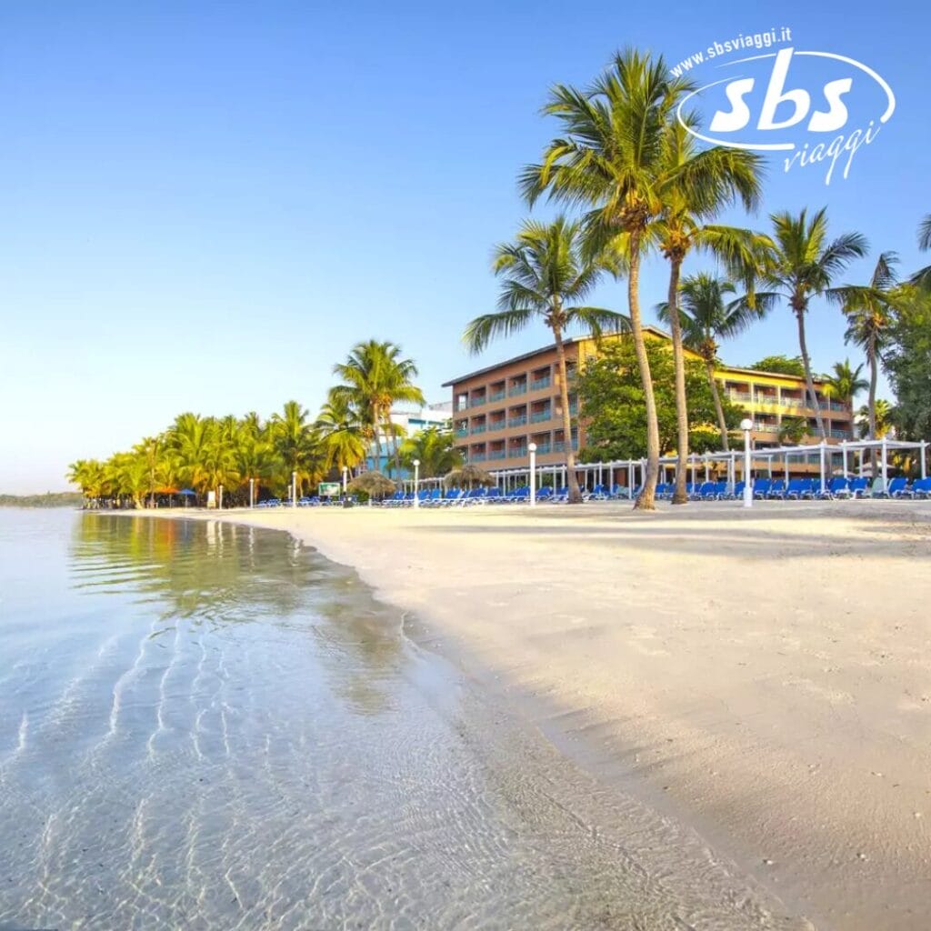 Resort sulla spiaggia con palme e lettini prendisole in riva all'acqua calma, dove l'atmosfera serena sembra quasi una bozza automatica. Un edificio a più piani si erge sullo sfondo sotto un cielo limpido.
