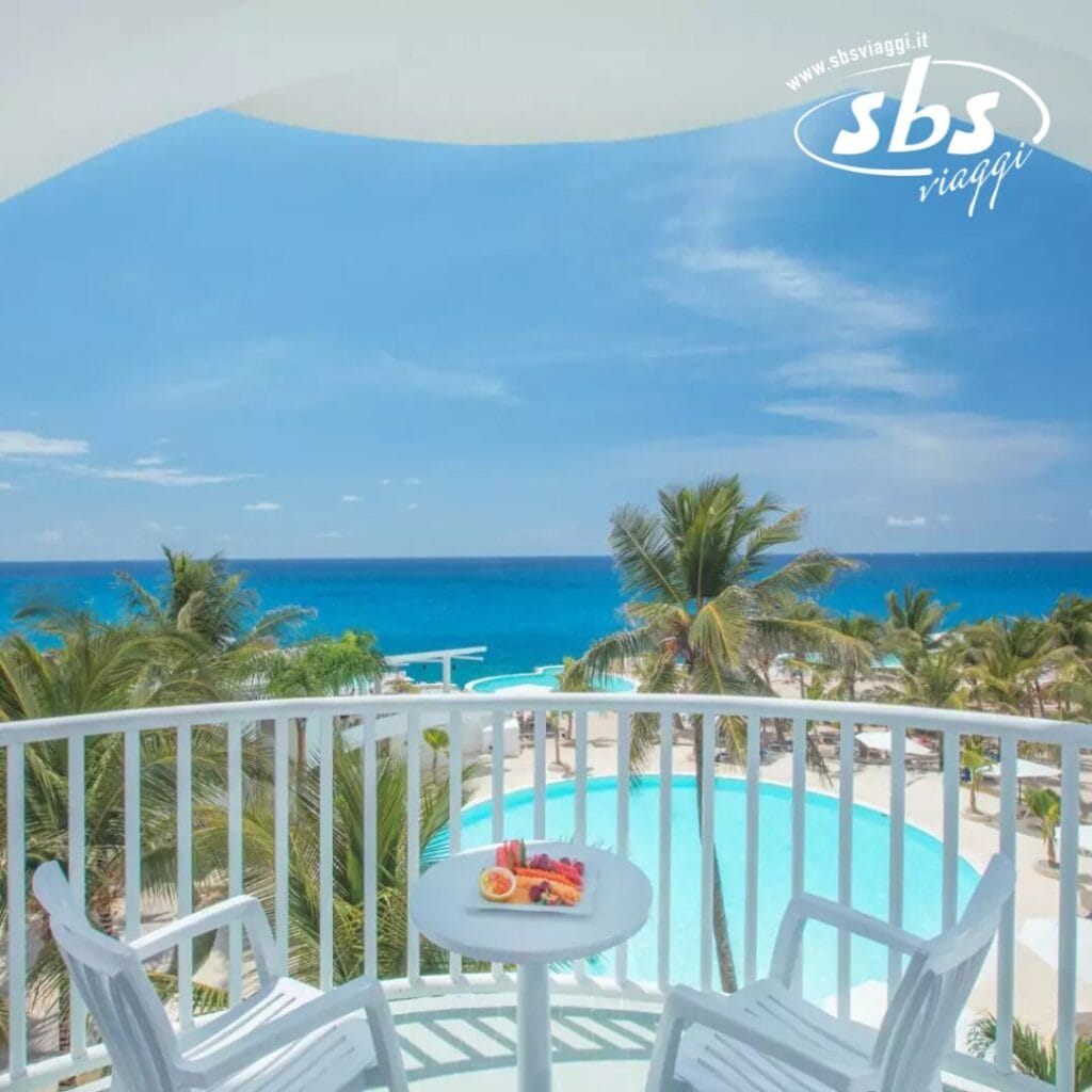 Dal balcone, godetevi la vista mozzafiato: una piscina tranquilla, palme ondeggianti e il vasto oceano sotto un cielo azzurro e limpido, il tutto incorniciato da due invitanti sedie e un grazioso tavolo in primo piano.