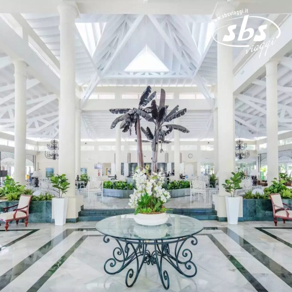 La spaziosa hall del Grand Bahia Principe presenta soffitti alti, pilastri bianchi e un tavolo centrale in vetro decorato con una pianta vivace. Le decorazioni con palme completano le eleganti aree salotto, creando un'atmosfera invitante che ricorda il fascino di El Portillo.