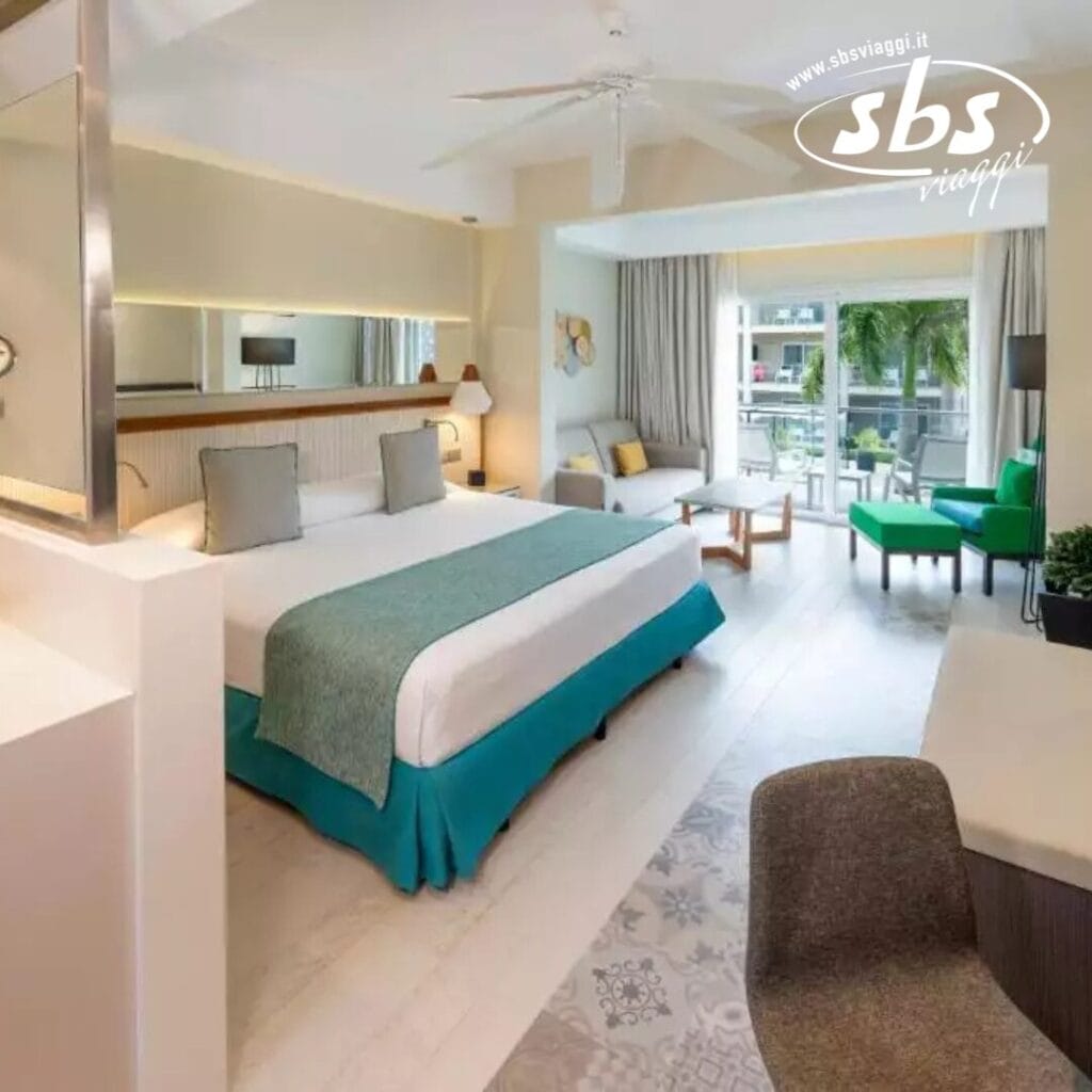 Una moderna camera d'albergo al Catalonia Royal, dotata di un letto grande, area soggiorno, scrivania e ventilatore a soffitto. Una finestra si affaccia sulle palme. Esclusivamente per ospiti di età superiore ai 18 anni a La Romana. Logo SBS Viaggi nell'angolo in alto a destra.