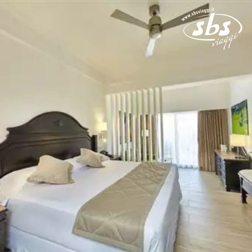 Provate il comfort di una spaziosa camera d'albergo al Riu Palace, dotata di un grande letto con cuscini morbidi, comodini e un ventilatore a soffitto. Una parete divisoria separa elegantemente la zona notte dalla porta scorrevole in vetro che si apre su un balcone con vista sulle viste mozzafiato di Punta Cana.