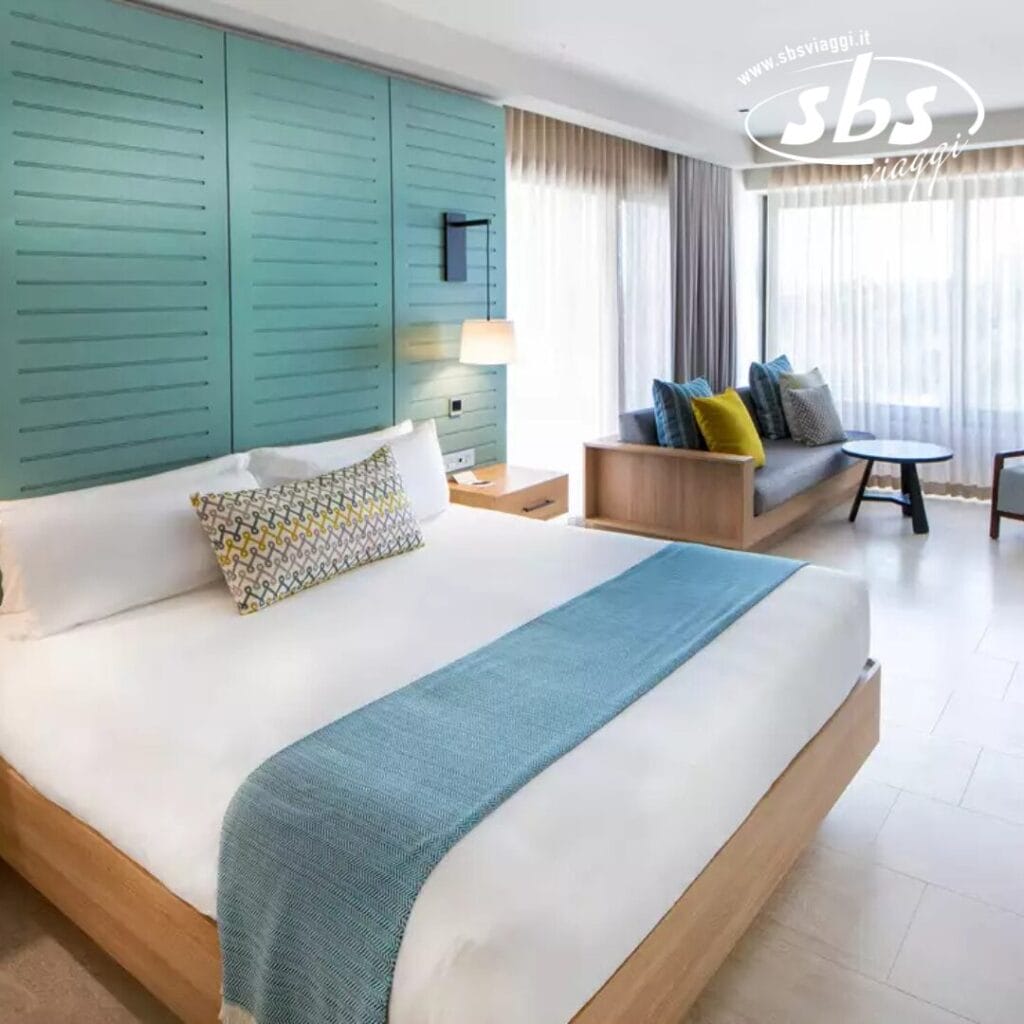 Moderna camera d'albergo con un grande letto con coperta blu, cuscino fantasia e illuminazione automatica Bozza. Godetevi una piccola area salotto e finestre dal pavimento al soffitto con tende trasparenti che lasciano entrare la giusta quantità di luce naturale.