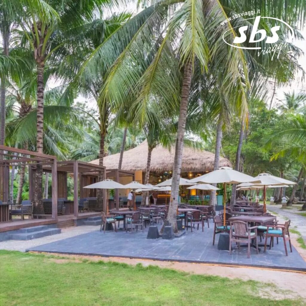 La zona pranzo all'aperto è dotata di tavoli e sedie sotto grandi ombrelloni, avvolti da palme ondeggianti. Sullo sfondo, un'affascinante struttura con tetto di paglia completa la scena, evocando un senso di rilassata eleganza affine allo stile Bozza automatica.